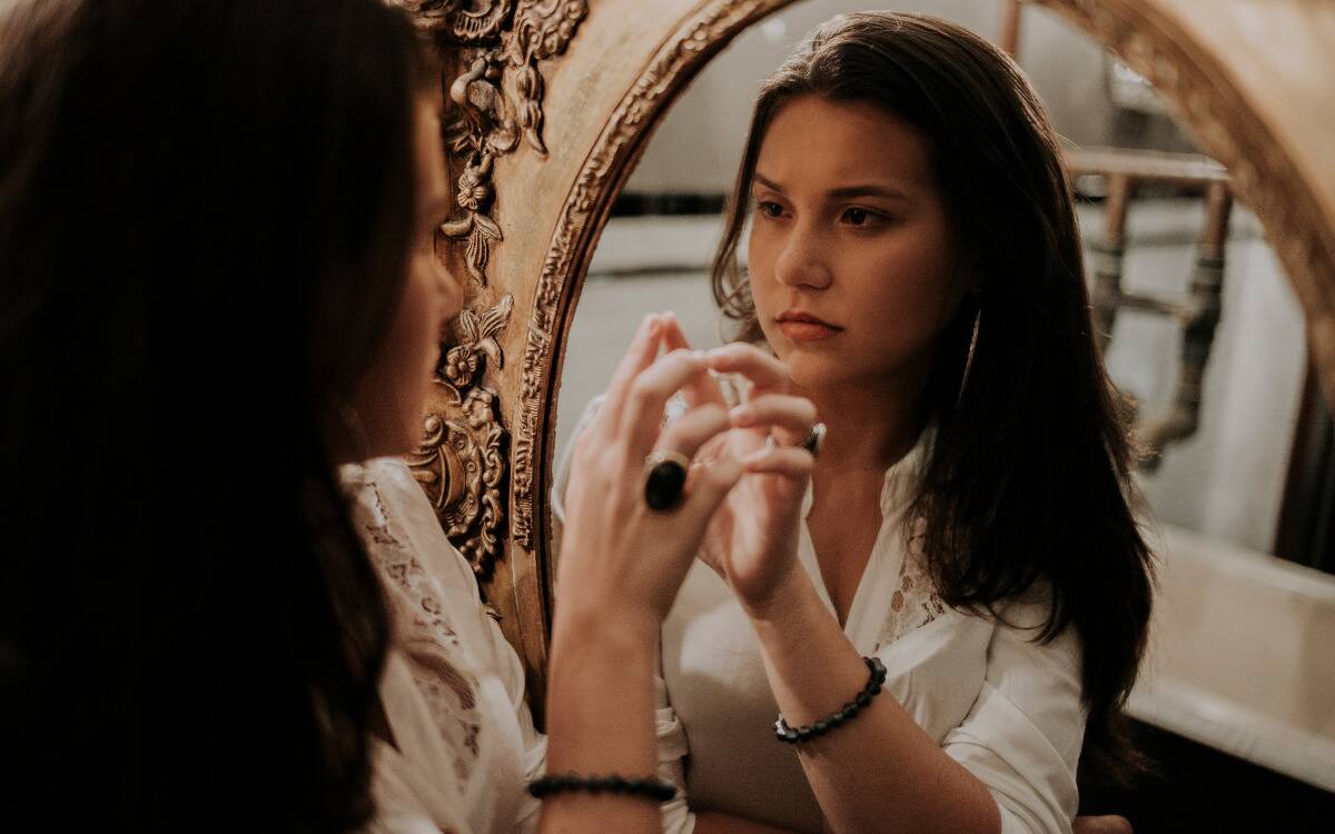 A woman looking thoughtfully into the mirror, rising a hand to touch her reflection.