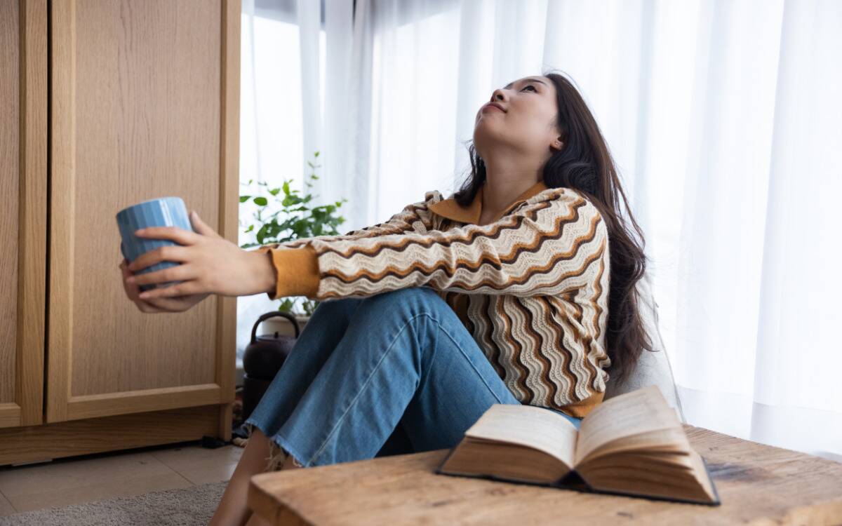 A woman sitting at home, a mug in hand, arms extended forward resting on her knees while she looks upwards.