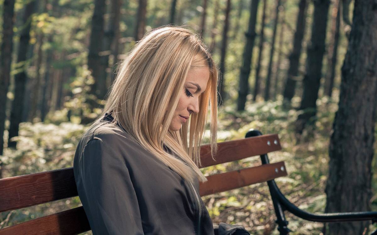 A woman sitting on a bench in a forest, looking down at her lap.