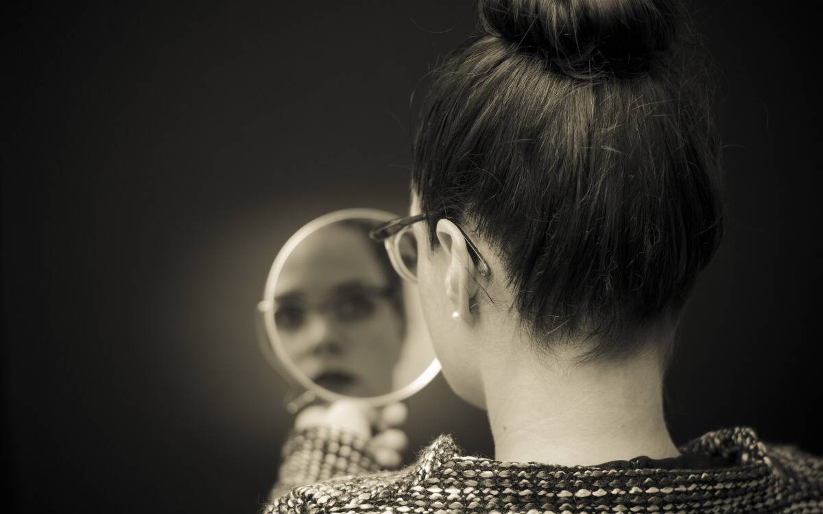 A greyscale image of a woman looking in a handheld mirror, shot from behind but her face visible in the reflection.