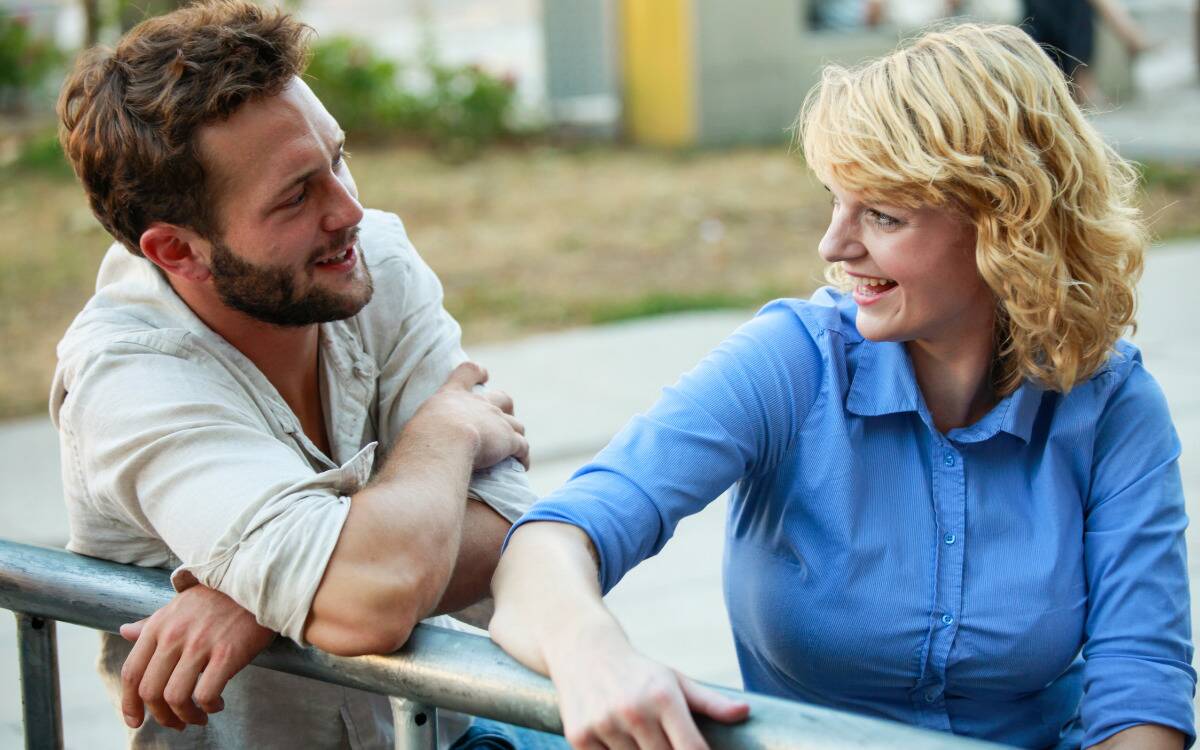 A man and a woman having a conversation on a bench outside, both smiling. 