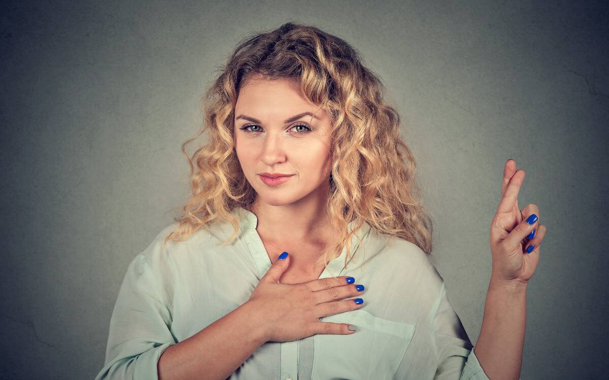 A woman looking to the camera with a suspicious expression, hand over her heart while also raising crossed fingers. 