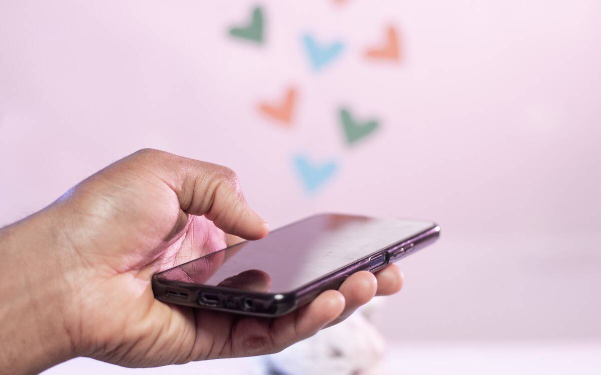 Someone holding out their phone against a blurred pink background with hearts. 
