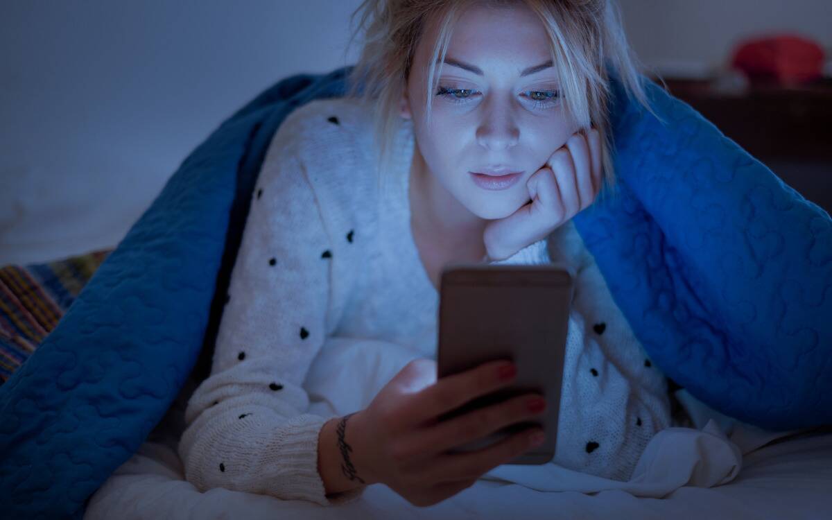 A woman looking at her phone in bed, lit up by the screen, under a blanket.