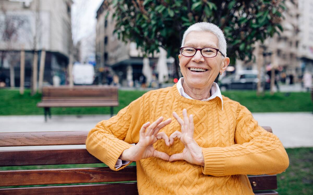 A woman in an orange sweater on a park bench, making a heart with her fingers in front of her chest.