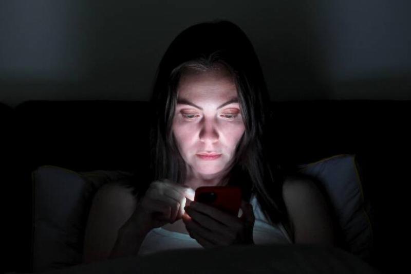 A woman awake in bed looking at her phone that's lighting up her face in the dark.
