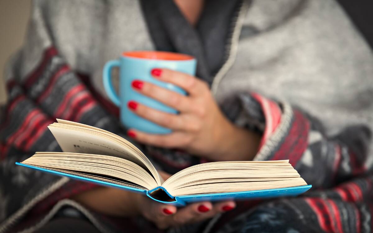 Someone reading a book, holding it open in one hand while the other holds a mug.