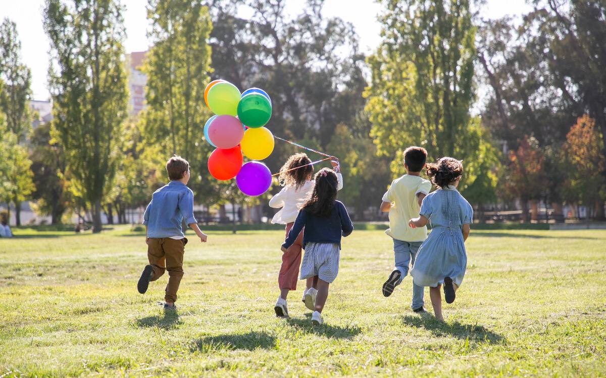 A group of kids running through a field, one of them holding a bundle of colorful balloons.