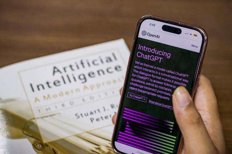 Someone holding up a phone with ChatGPTs home screen atop a book about AI.