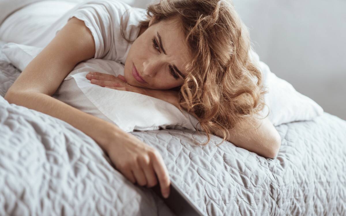 A woman looking sad as she lays in bed, phone in hand.
