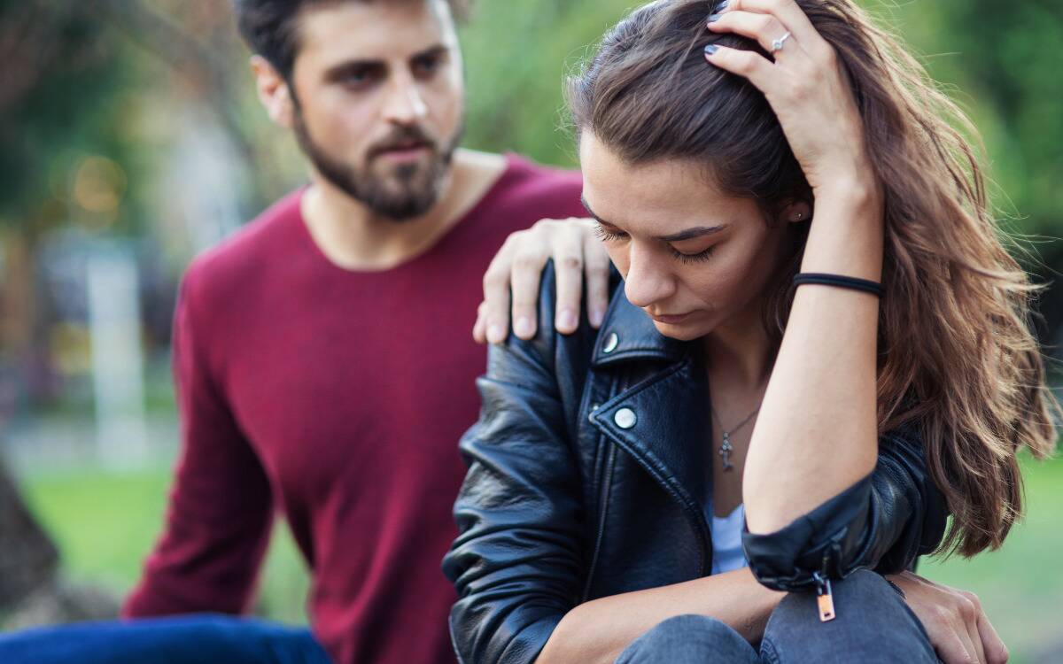 A couple fighting, the woman with a hand in her hair as she looks sad while the man insistently puts a hand on her shoulder.