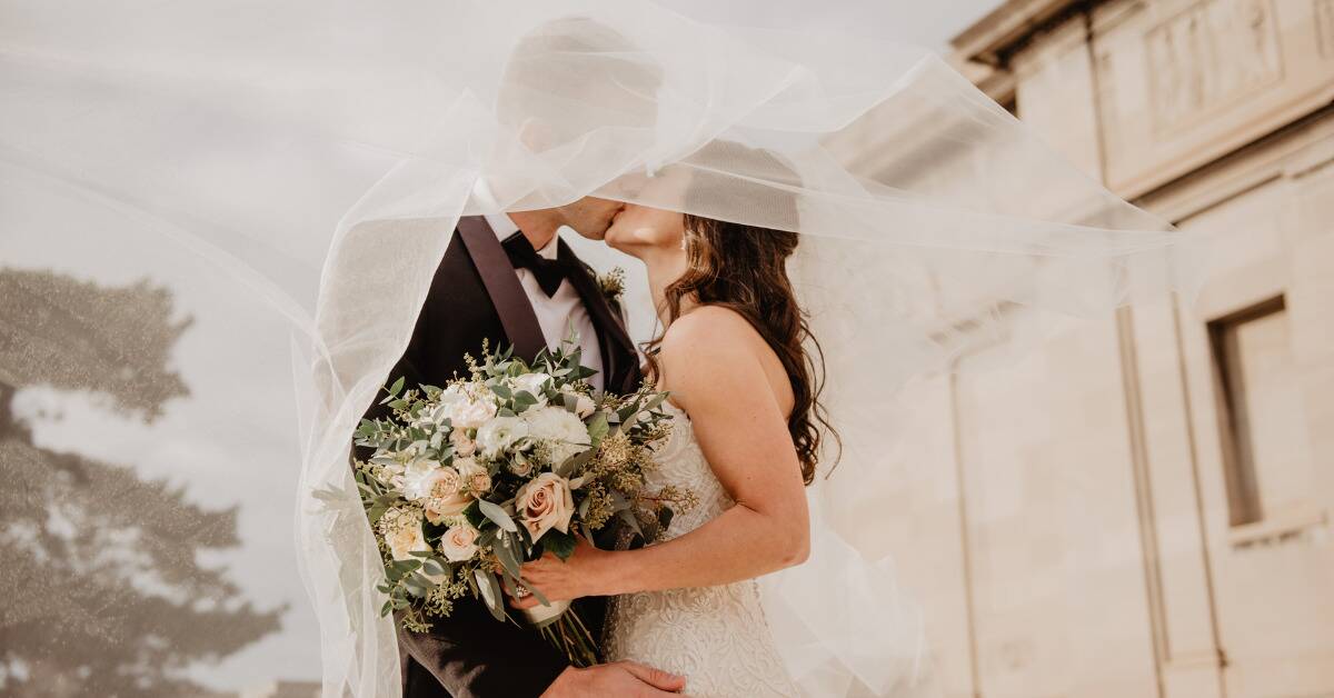 A newly married couple kissing under the bride's billowing veil.