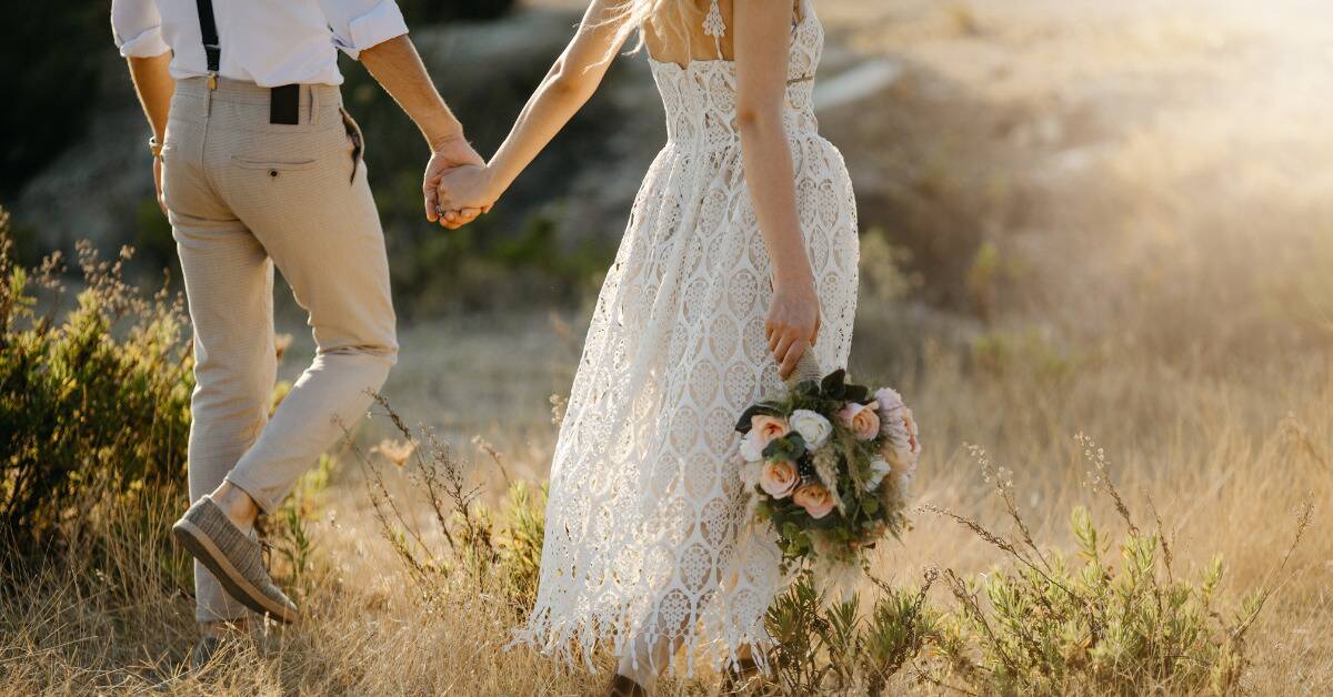 A woman in her wedding dress, holding her bouquet down at her side, walking through some tall grass.