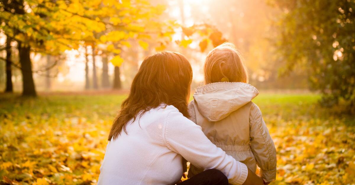 A woman kneeling down next to her daughter in the park, looking at the autumn yellow leaves.