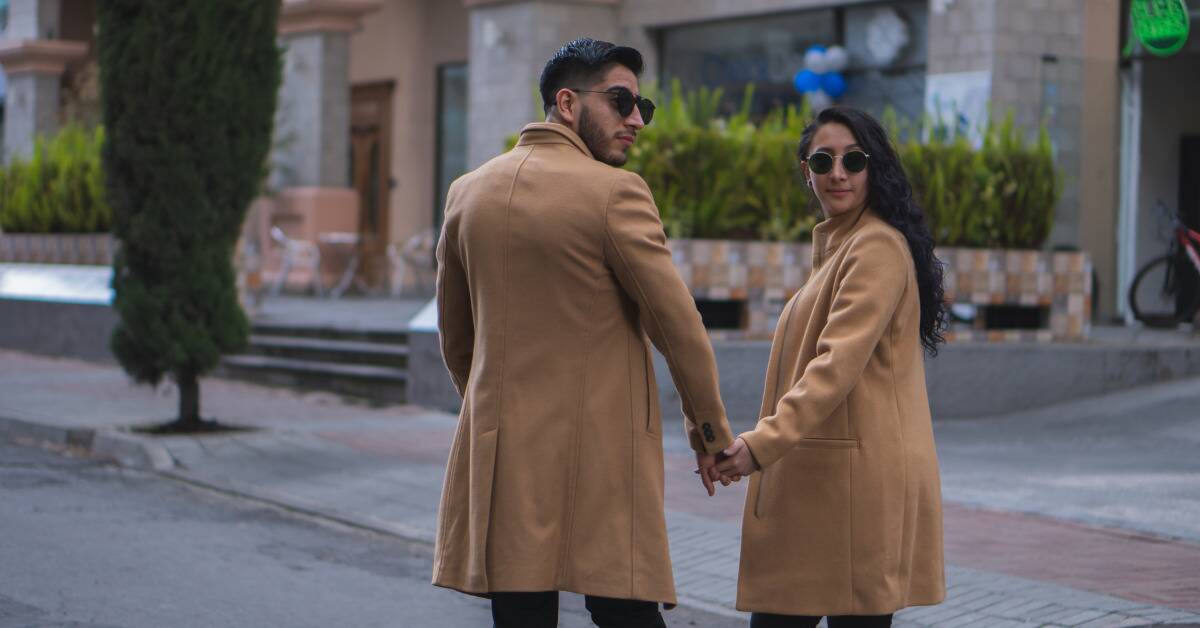 A couple holding hands, both looking over their shoulder, wearing matching tan coats and round sunglasses.