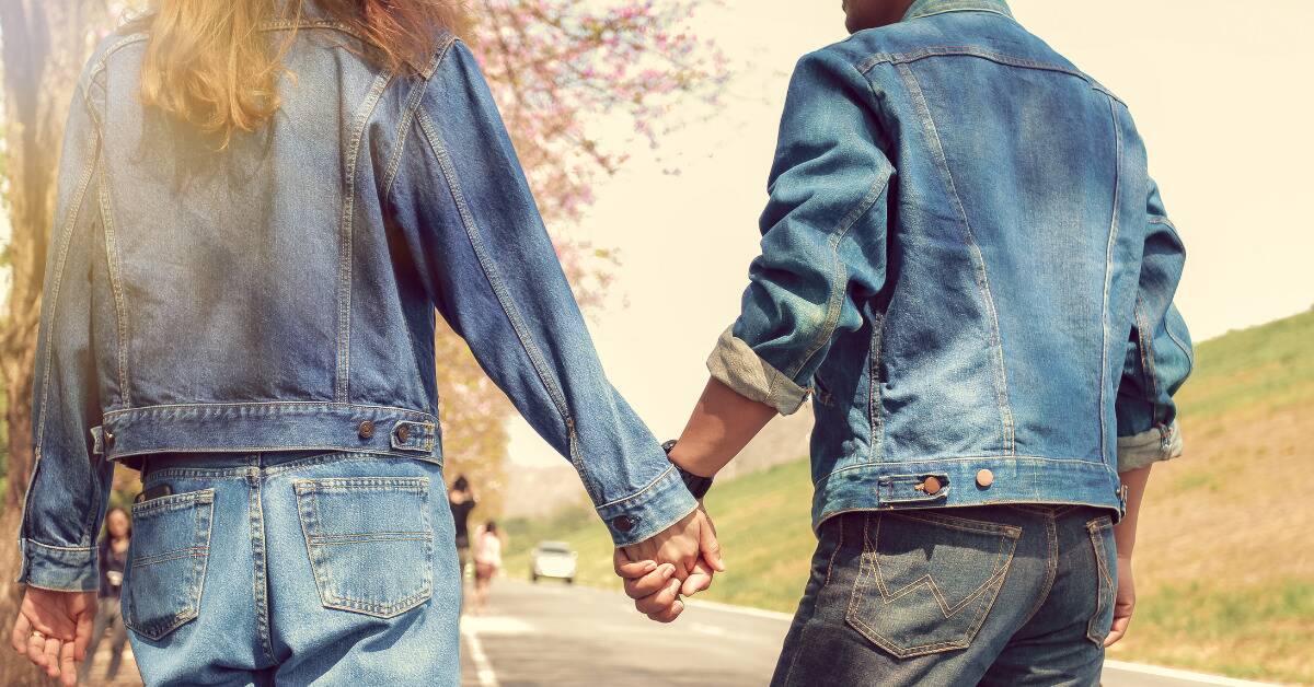 Two people in denim pants and jackets holding hands as they walk.