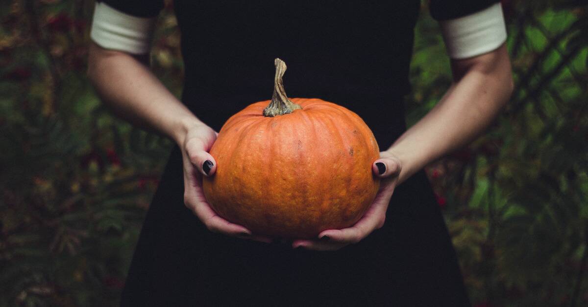 Someone in a black dress and with black painted nails holding a small pumpkin.