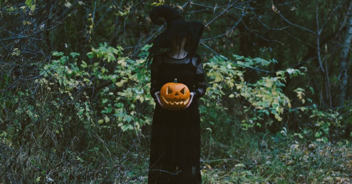 A woman in a witch outfit standing outside in the greenery, holding a jack-o-lantern.