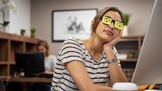 A woman asleep at work, eyes drawn on post it notes covering her glasses, a parody of trying to appear awake.