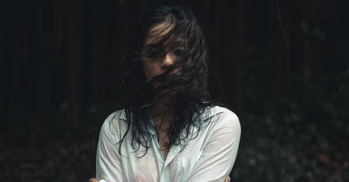 A woman standing outside, appearing to be caught in a rain storm, her hair and clothes wet while the wind blows her hair into her face.