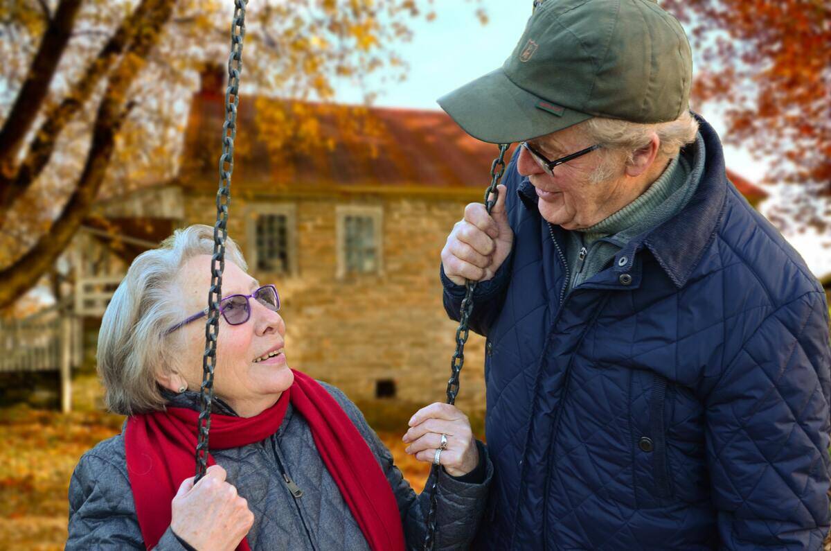 An older woman sitting on a park swing while an older man speaks to her, both are smiling.