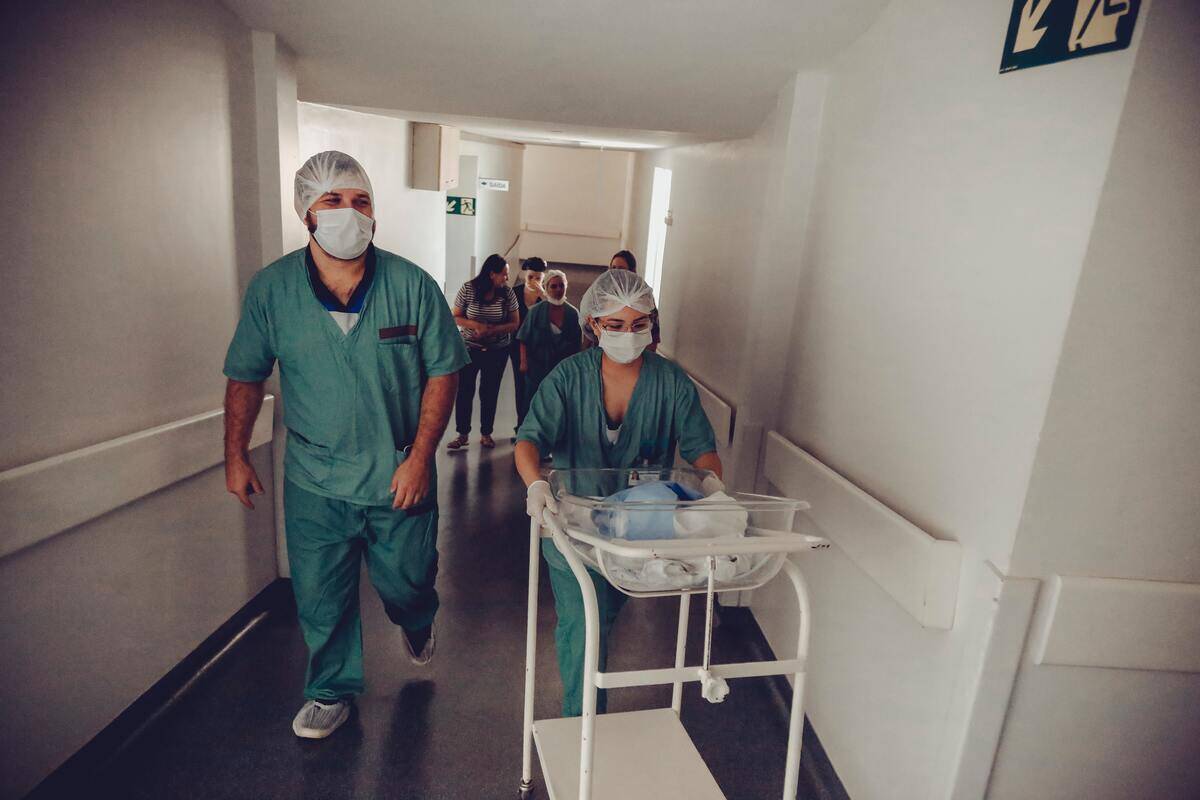 Two nurses walking through a hospital hallway, one pushing a cart with a baby in it.