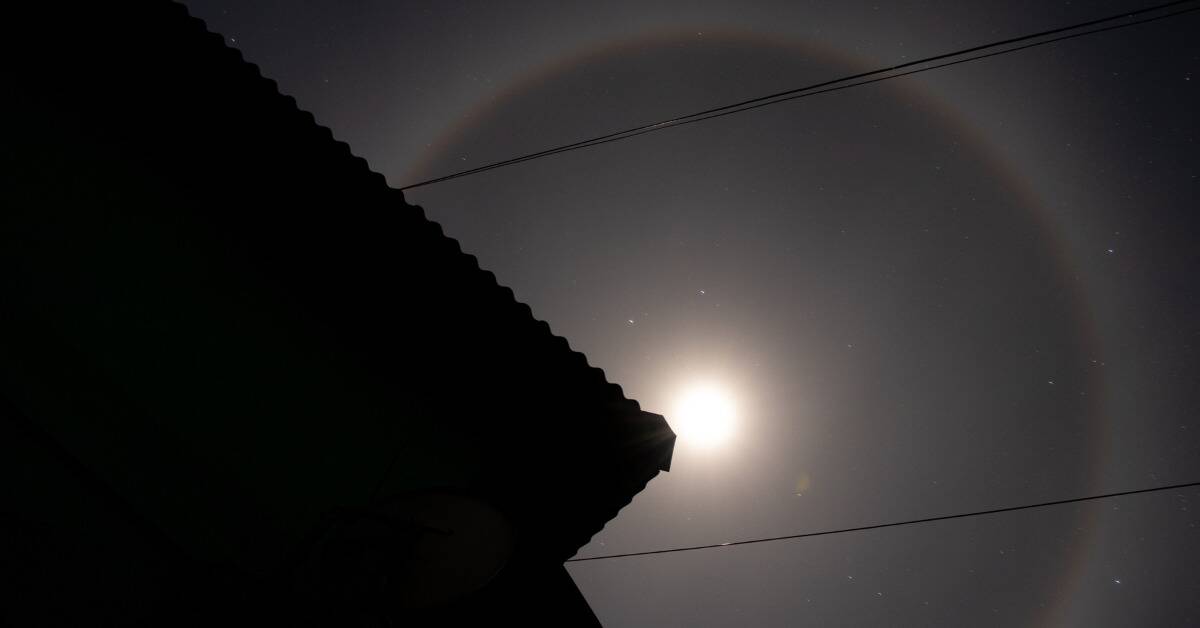 A lunar halo with the silhouette of a home in front of it, a corner of the roof pointed at the center.