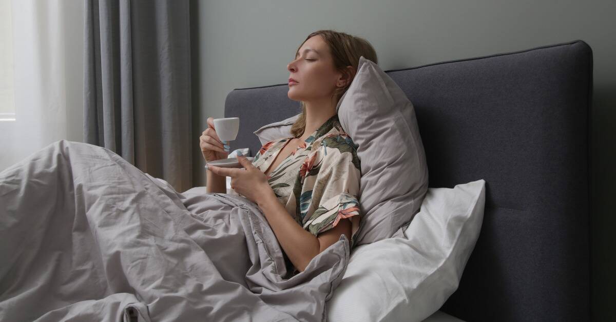 A woman sitting in bed, holding a teacup, her eyes closed.