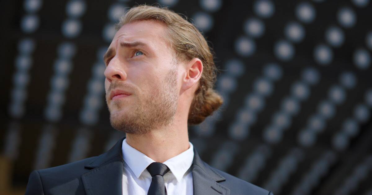 A man in a suit and long hair tied back, looking off to the side, observing.