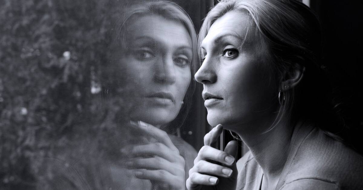 A greyscale image of a woman looking out a window in which her reflection is visible.