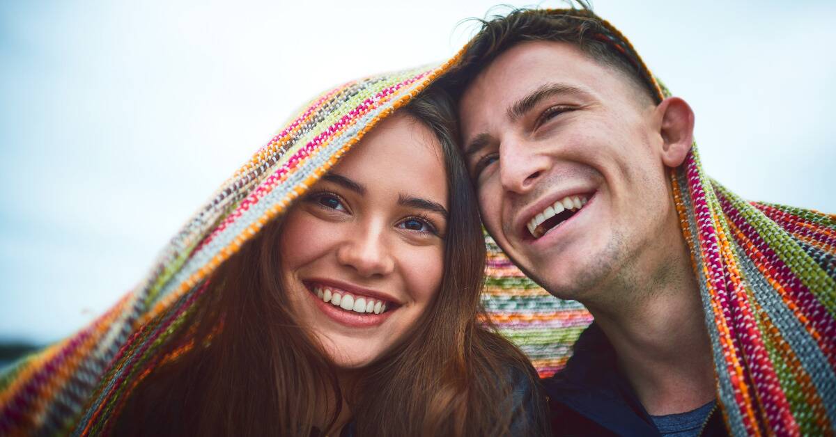 A couple smiling as they lean on each other under a blanket.