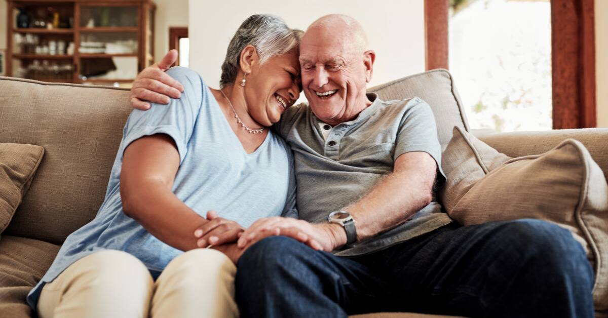 An older couple smiling as they embrace on the couch.