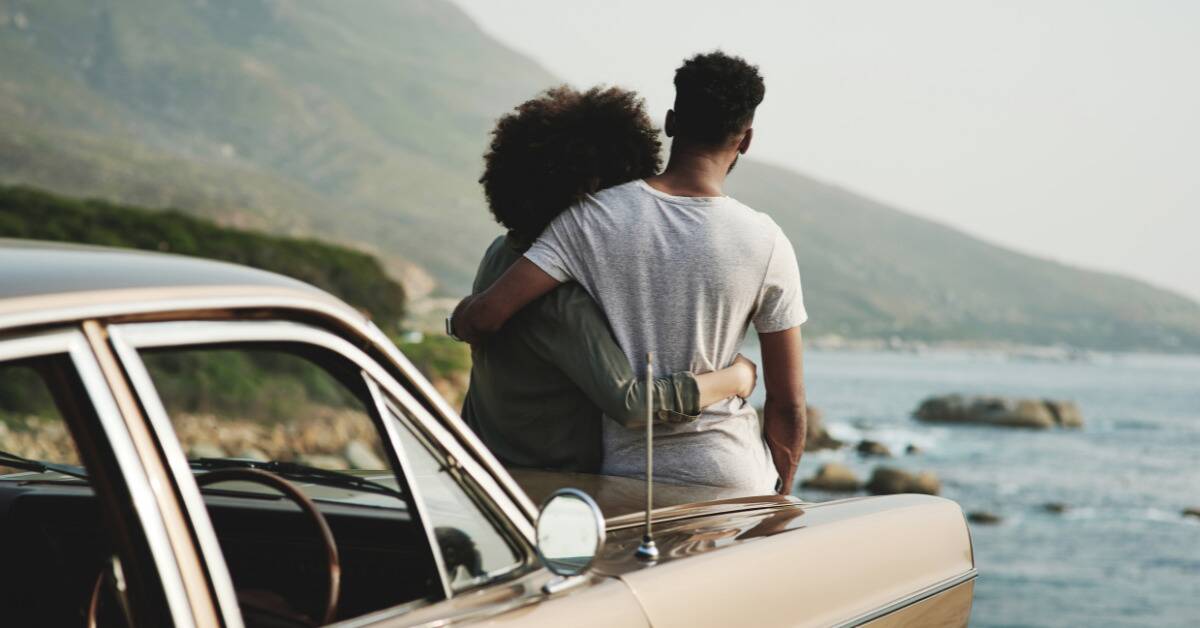 A couple sitting on the hood of a car overlooking the water, arms around one another.