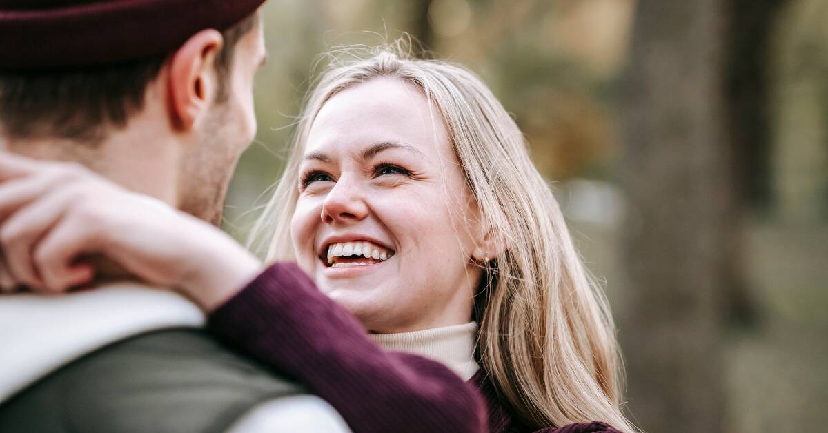 A couple standing with their arms around each other, shot over the man's shoulder, focusing on the woman's smiling face.