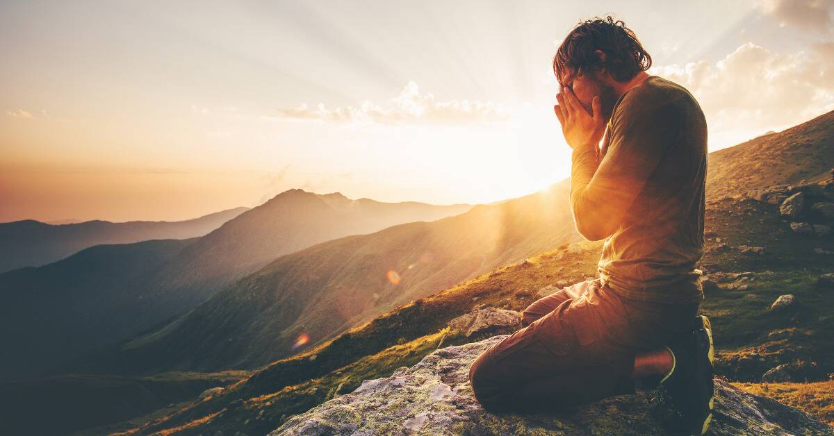 A man kneeling on a mountain range, head in his hands, the sun shining behind him.