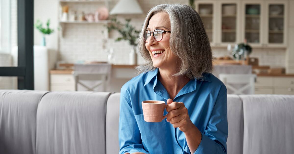 An older woman smiling, sitting on a couch as she drinks coffee.