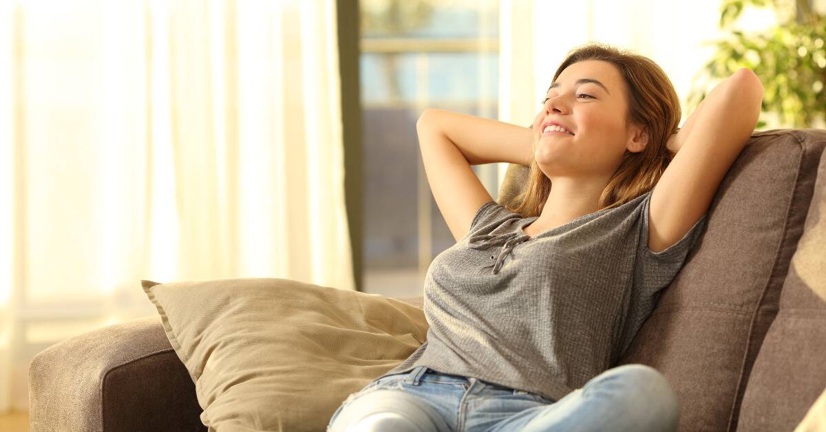 A woman sitting on her couch, hands behind her head, smiling as she relaxes.