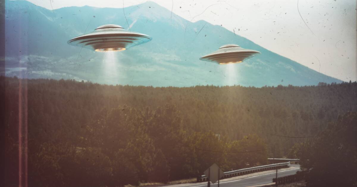 A grainy photo of two saucer UFOs hovering above a forest-lined road.