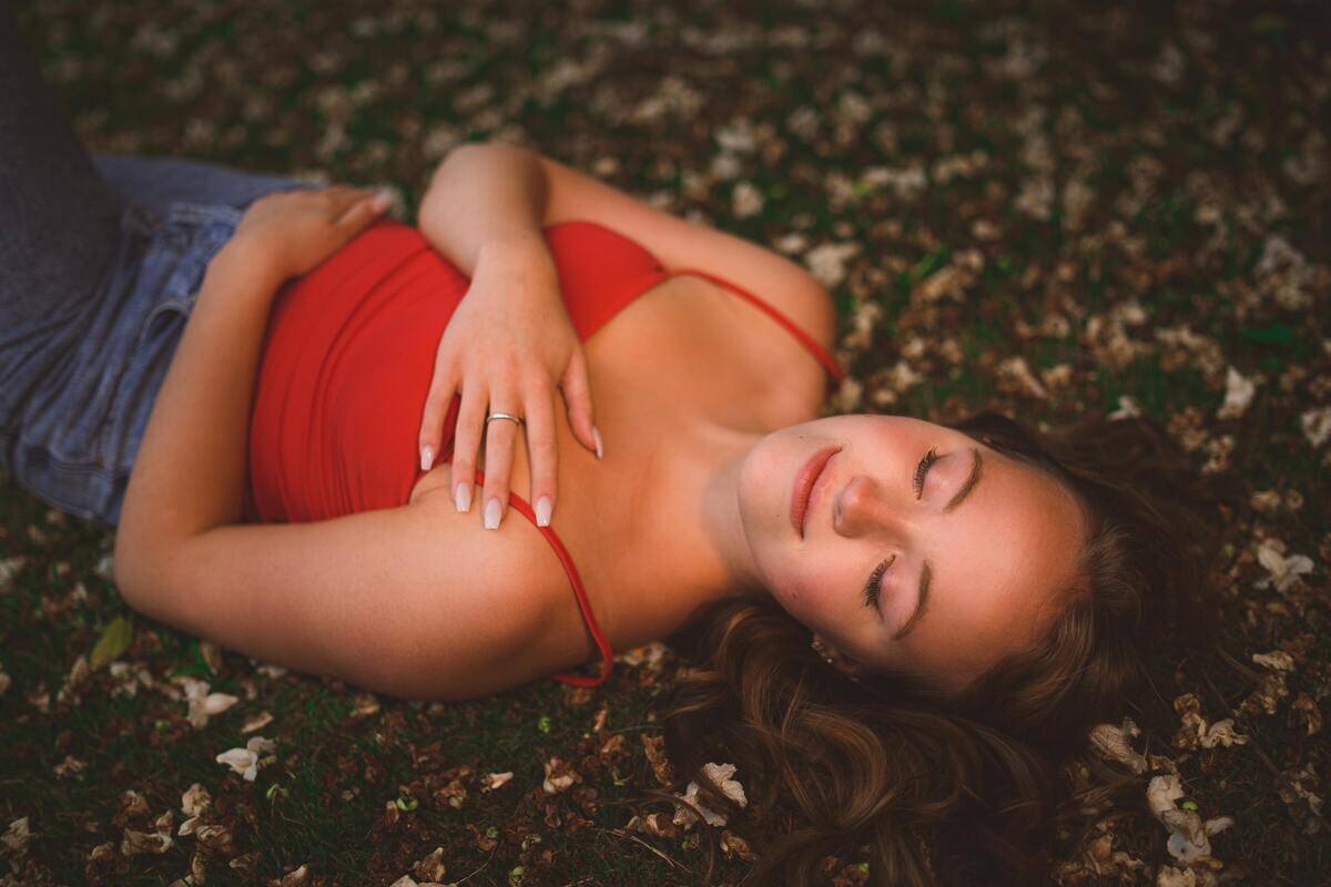 A woman laying on the grass outside, arms wrapped around herself, looking serene.