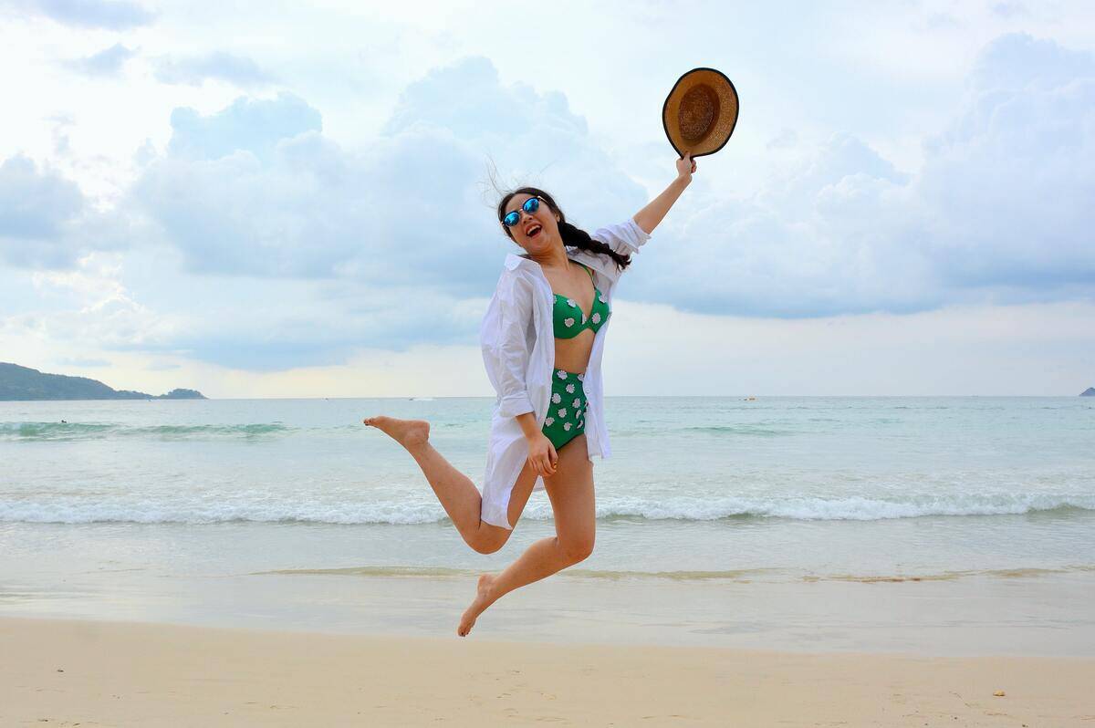 A woman on the beach jumping as she smiles, holding her hat up in the air.
