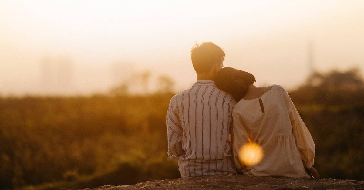 A couple sitting at sunset, the woman resting her head on the man's shoulder.