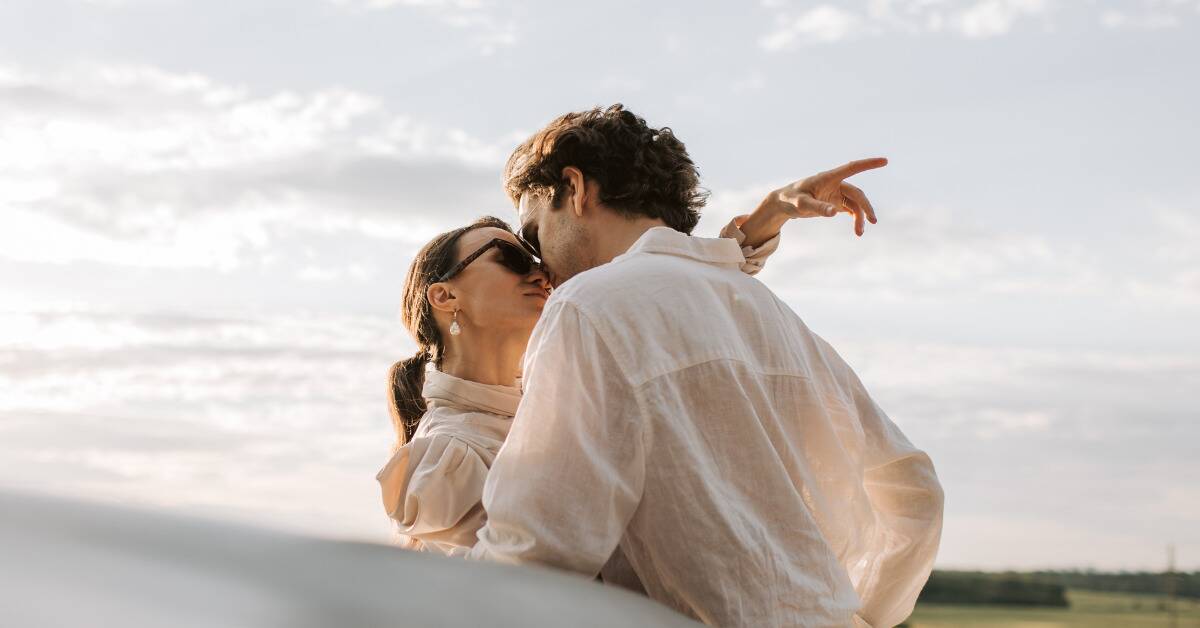 A couple embracing outside with their faces close together in an almost-kiss, the woman's hand loosely pointing.