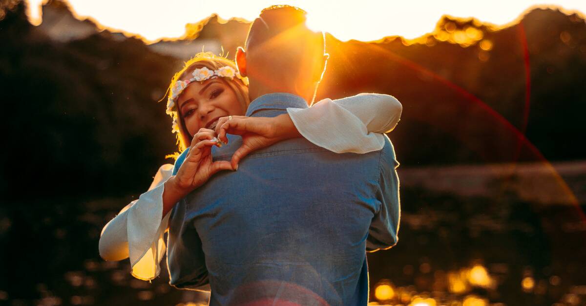 A man hugging his girlfriend, his back to the camera, while the woman smiles and makes a heart with her hands on the back of his shoulder.