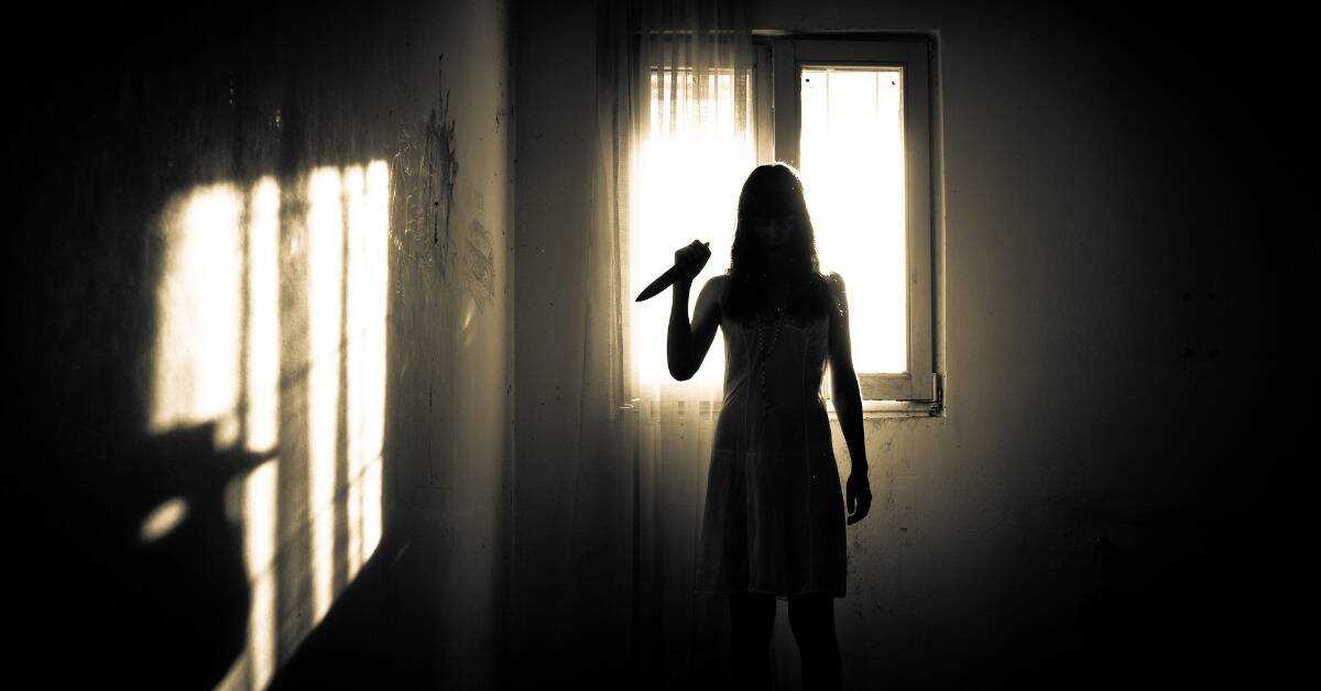 A silhouette of a woman holding up a knife as she stands in front of a window.