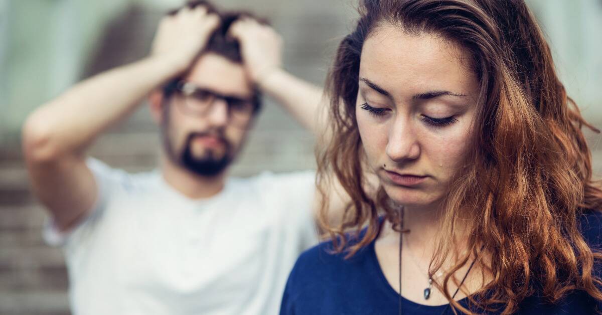 A woman looking sad while a man, out of focus in the background, has his hands in his hair.
