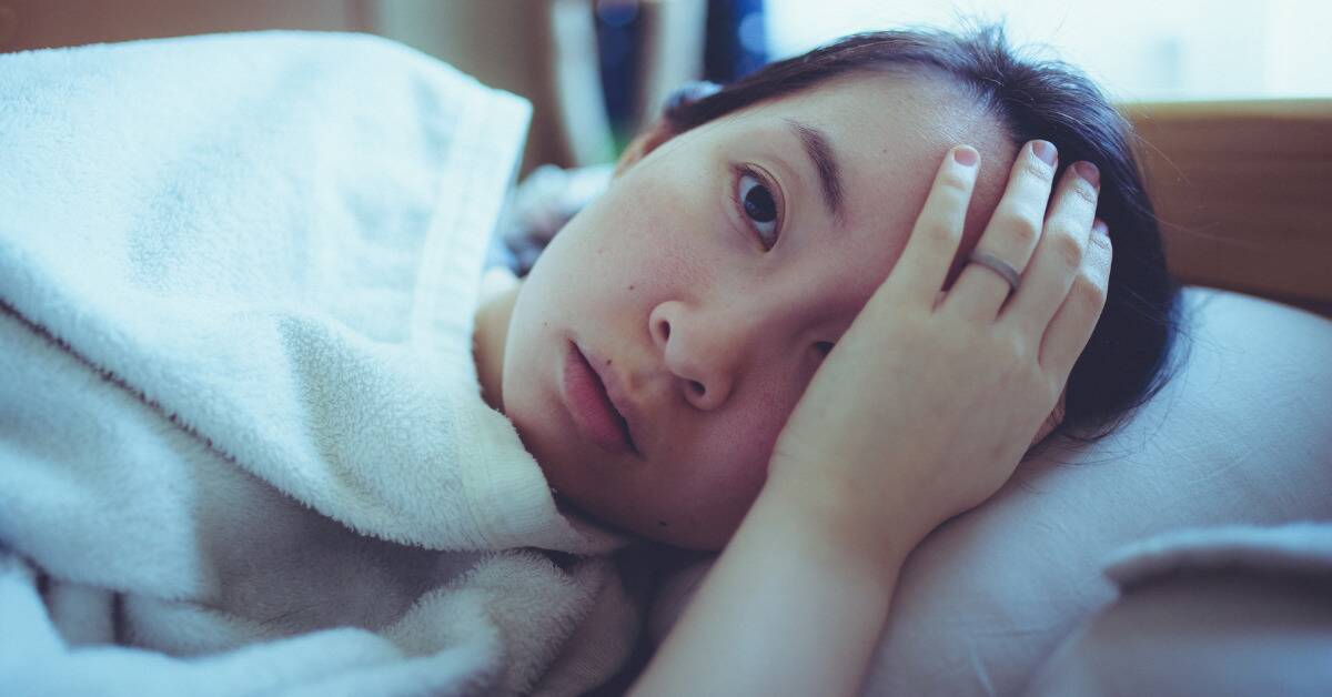 A young woman in bed, a hand covering one eye.