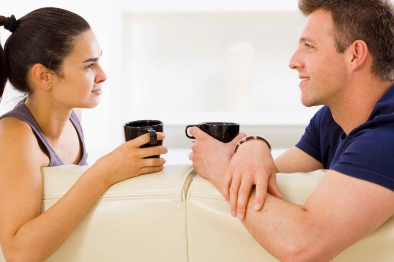 Couple talking to each other, sitting on a couch, and holding black mugs.