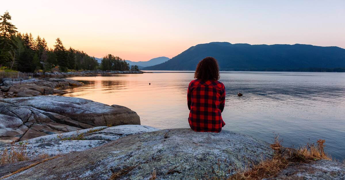 A woman sitting on a rock by a lake, looking outward.