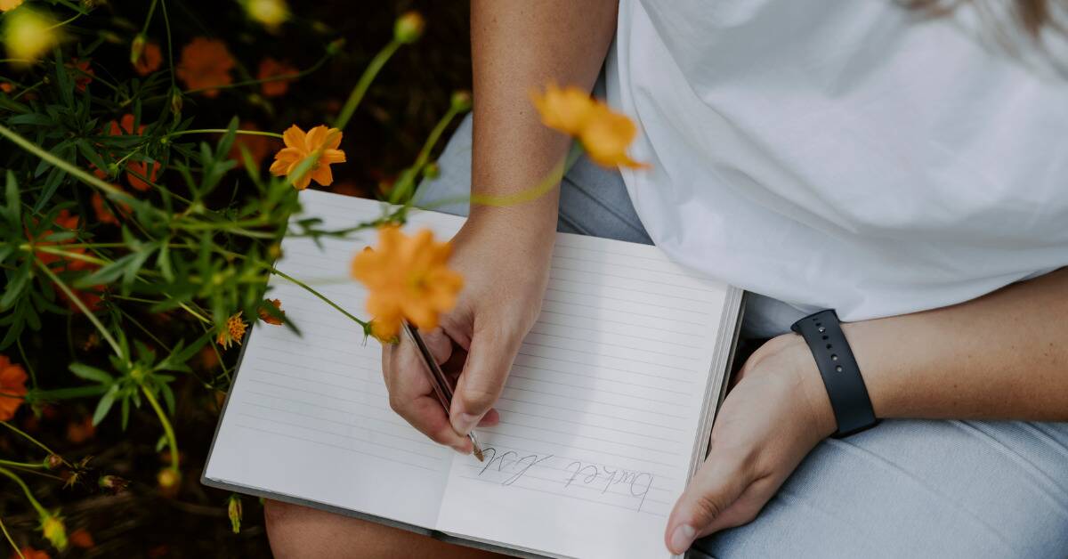 Someone writing a bucket list in a journal by a bush of yellow flowers.