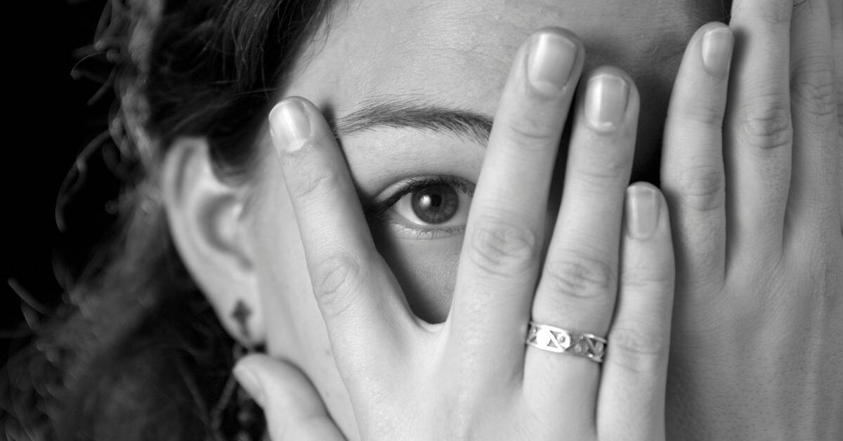 A greyscale image of a woman peeking through her fingers that are covering her face.
